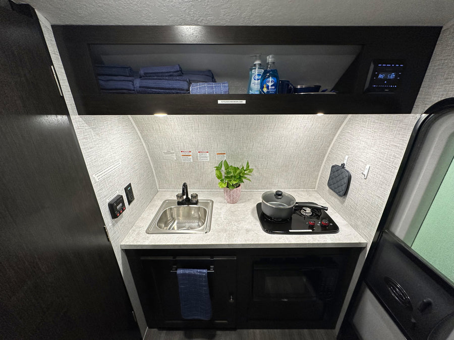 Inside an RV rental, a compact kitchen features a sink, two-burner stove, microwave, and control panel. Ready for your adventure!