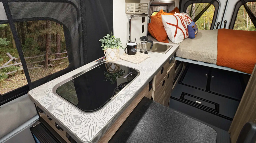 Cozy RV rental interior: kitchen with map-pattern counter, induction cooktop, sink, and comfy bed. Forest view through mesh window.