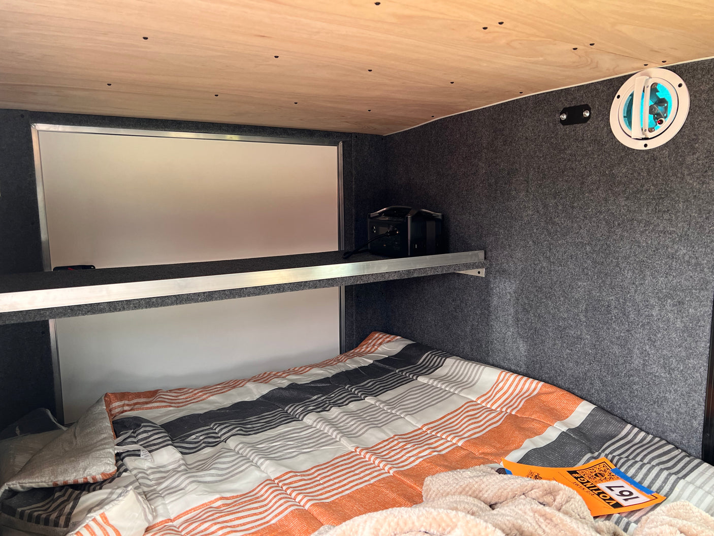 Cozy RV rental interior with a striped bed, overhead shelf holding a power station, and side wall vent for a comfortable sleep setup.