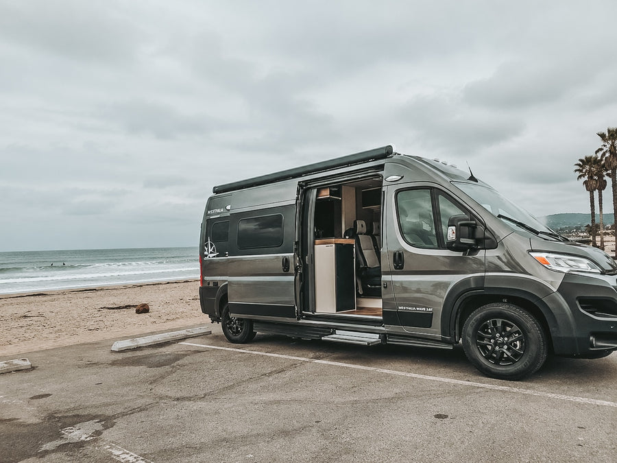 Grey Westfalia camper van rv rental parked by a sandy beach with surfers. Ideal for oceanfront adventures.|Open doors of this rv rental camper van show a cozy interior at a cloudy beach. Your next travel adventure awaits by the ocean!|Experience an rv rental! This black Westfalia camper van with open rear doors sits on wet pavement at dusk