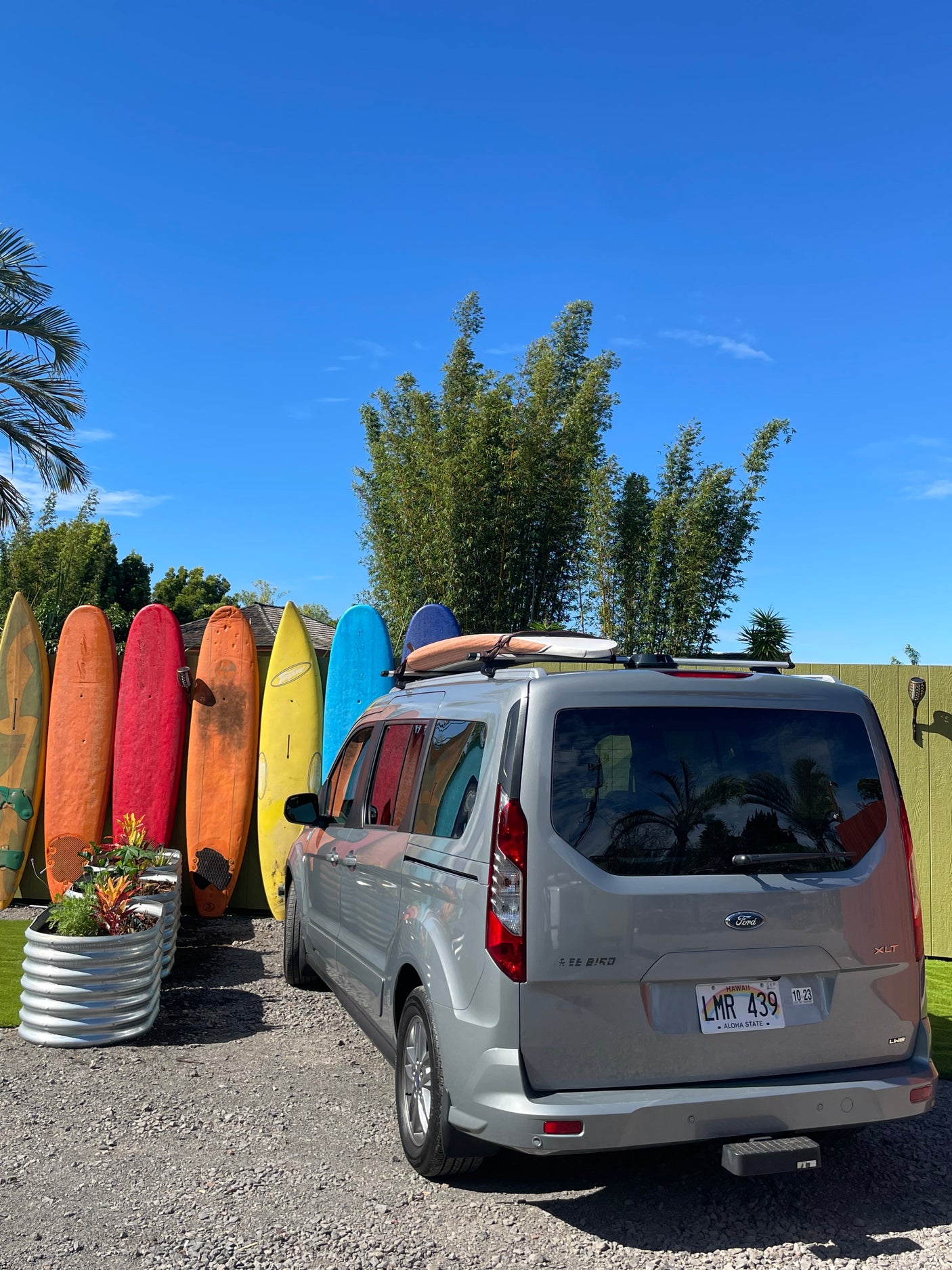 Explore Hawaii with this RV rental: A grey van with surfboards on its roof, parked near a vibrant display of colorful surfboards.