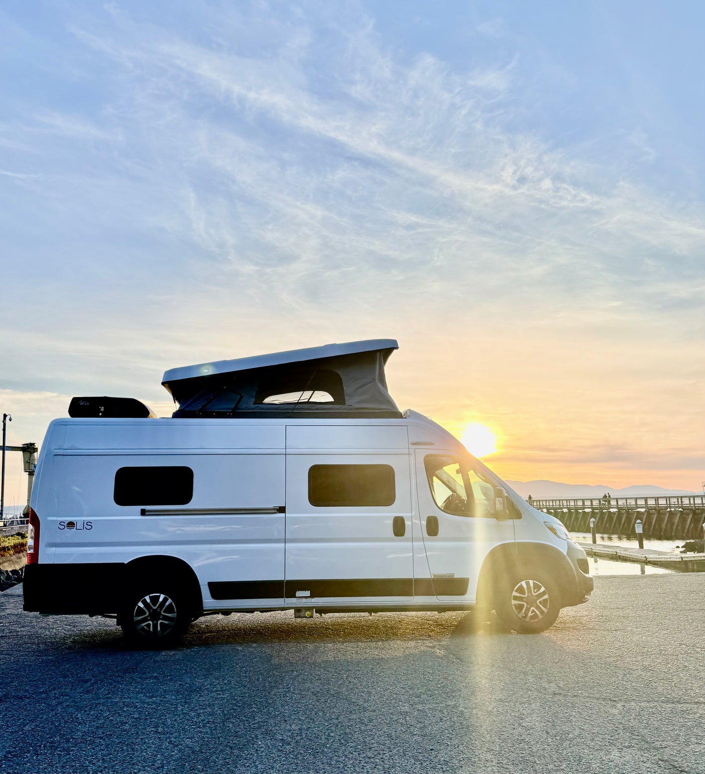 White Winnebago Solis camper van with pop-top open at sunset by a pier, perfect for an RV rental adventure.