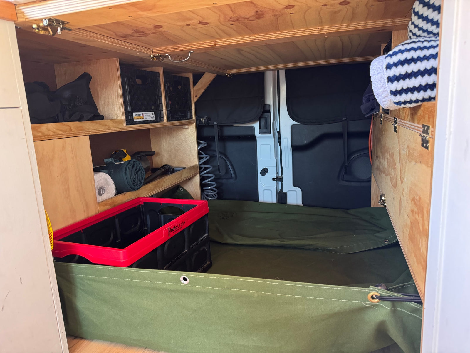 RV rental camper van interior storage: organized wooden shelves, crates, rolled gear, and a green canvas floor bin.