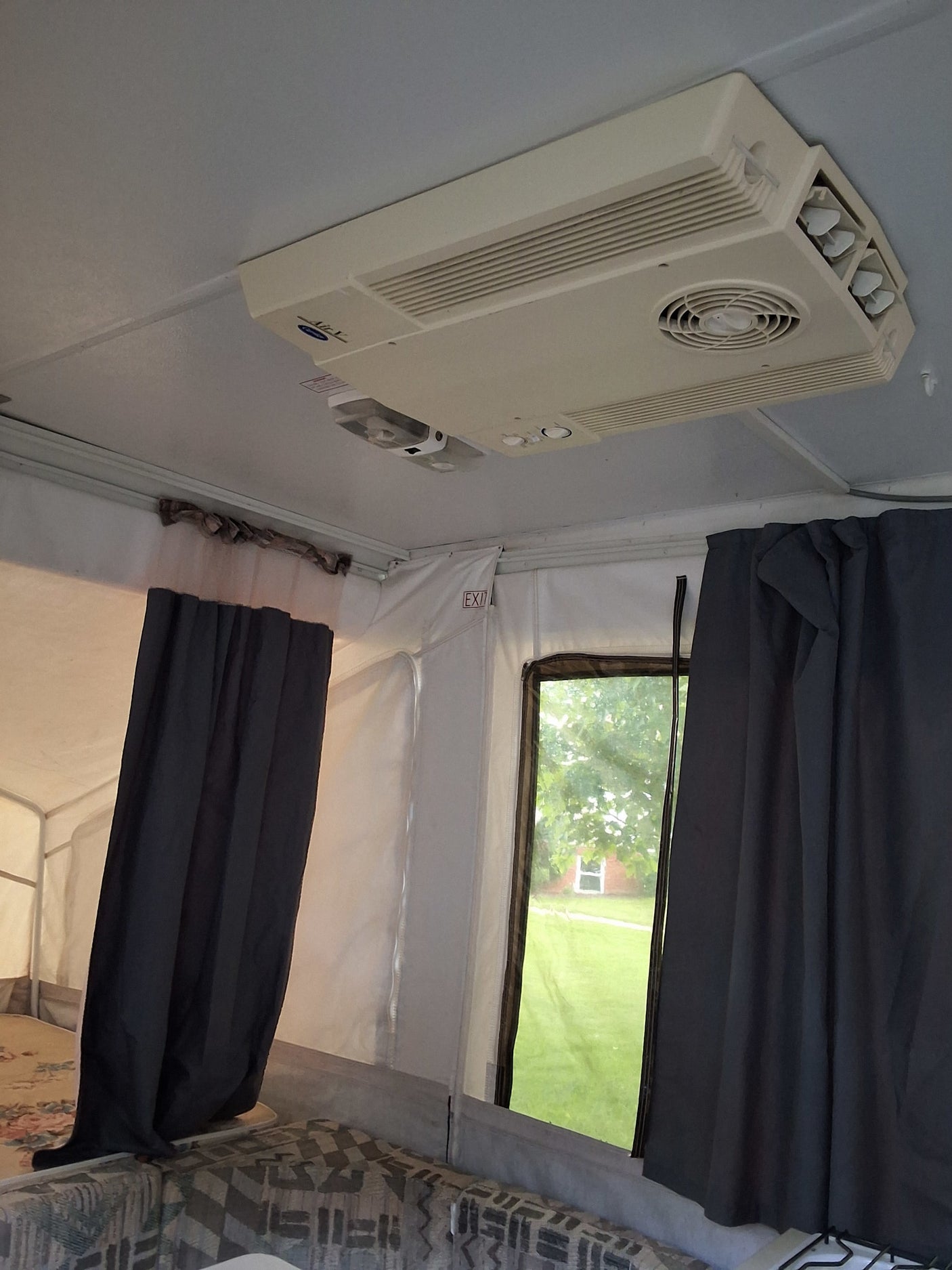 RV rental pop-up camper interior, featuring ceiling AC, dark curtains, window view, and an EXIT sign.
