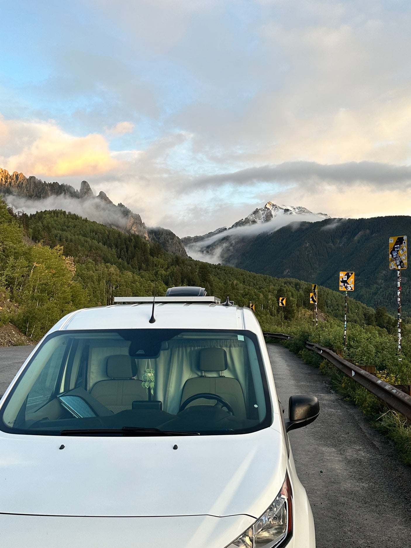 A white RV rental van parked on a scenic mountain road with misty, forested peaks and sticker-covered signs.