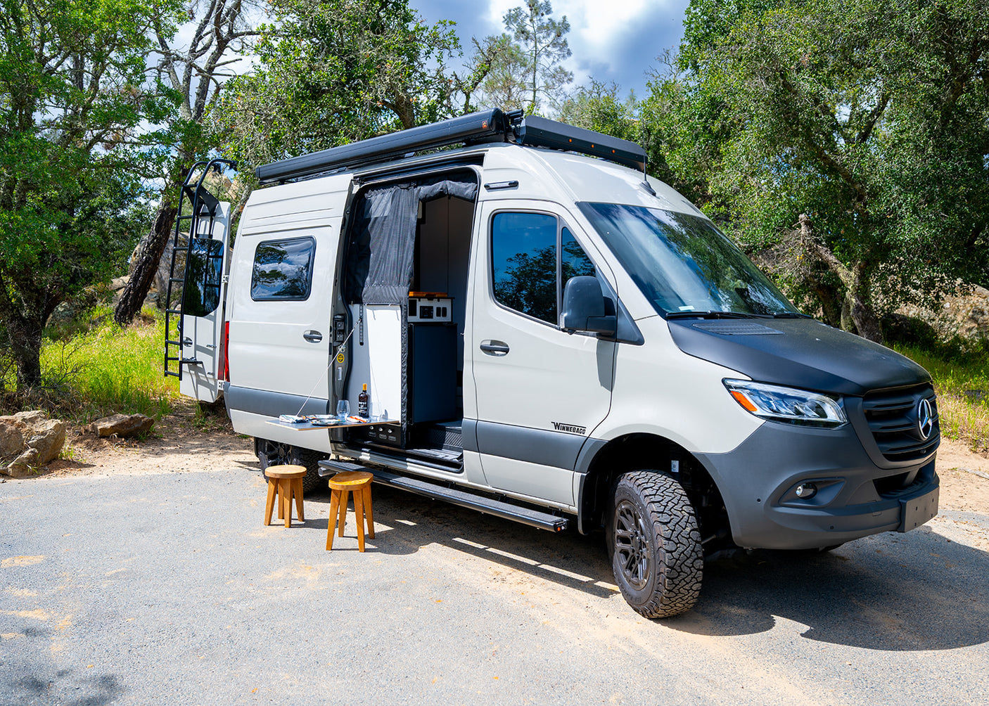 Winnebago RV rental camper van parked in nature. Side door open, revealing interior, with outdoor table, drinks & stools.