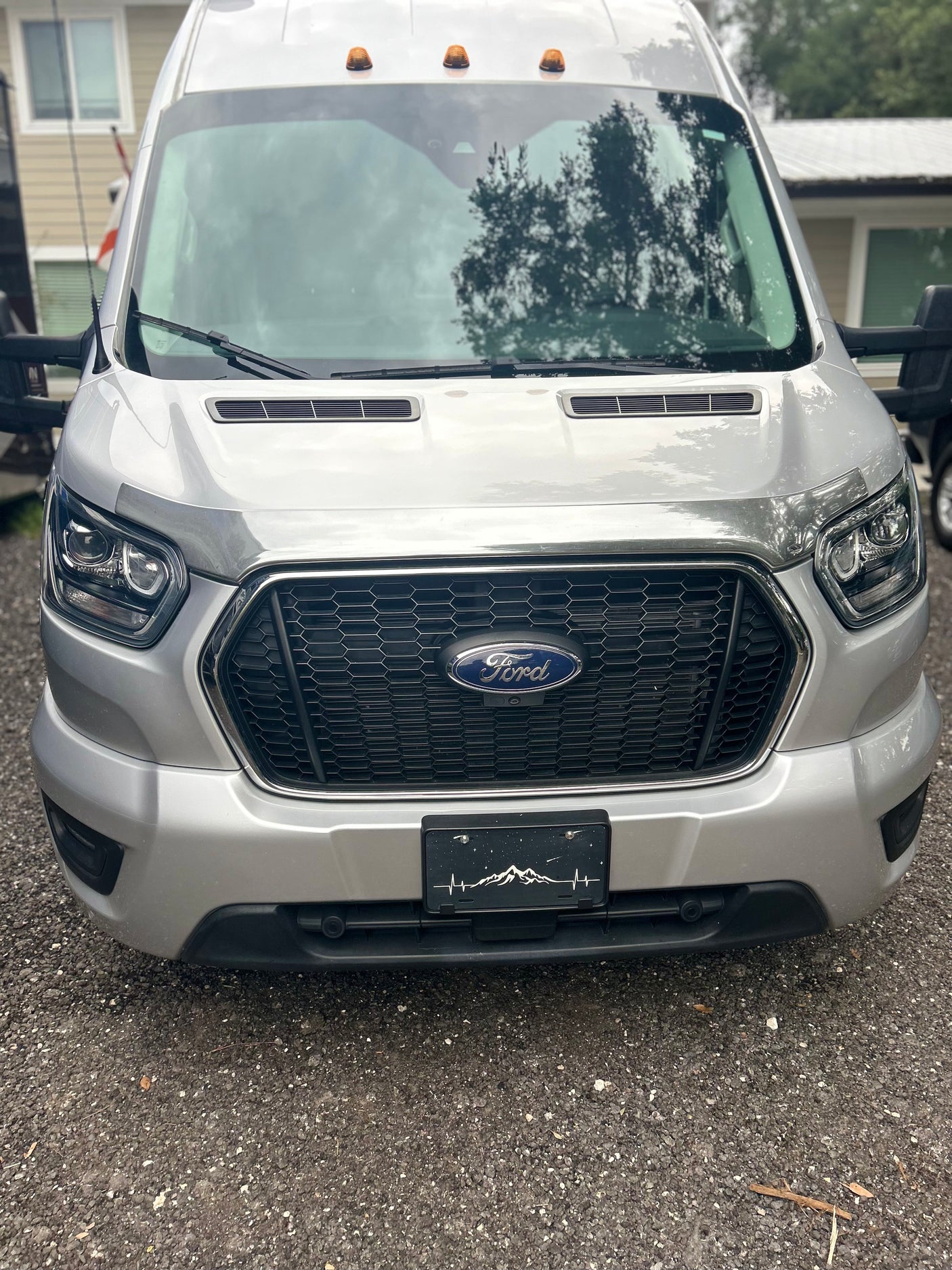 Silver Ford Transit van, likely an RV rental, shown from the front with a unique mountain-themed license plate frame.