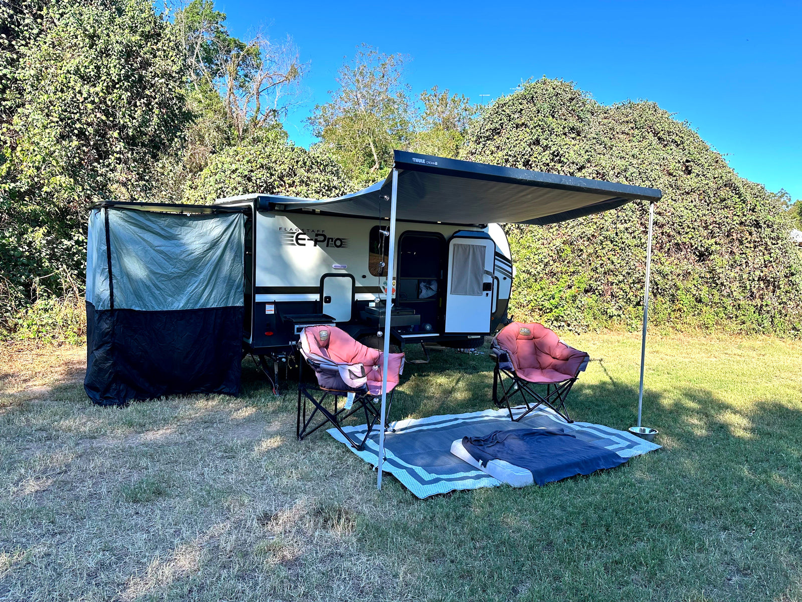 An RV rental, a Flagstaff E-Pro, is set up on a grassy site with its awning, side tent, chairs, and a mat under blue sky.
