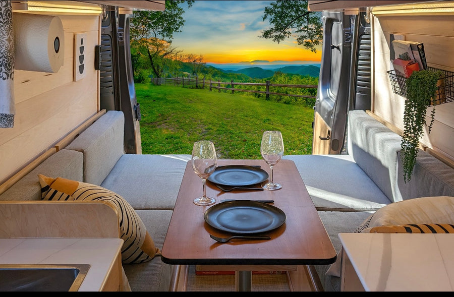 RV rental: Inside a cozy campervan, a table for two with wine glasses faces a stunning mountain sunset vista.