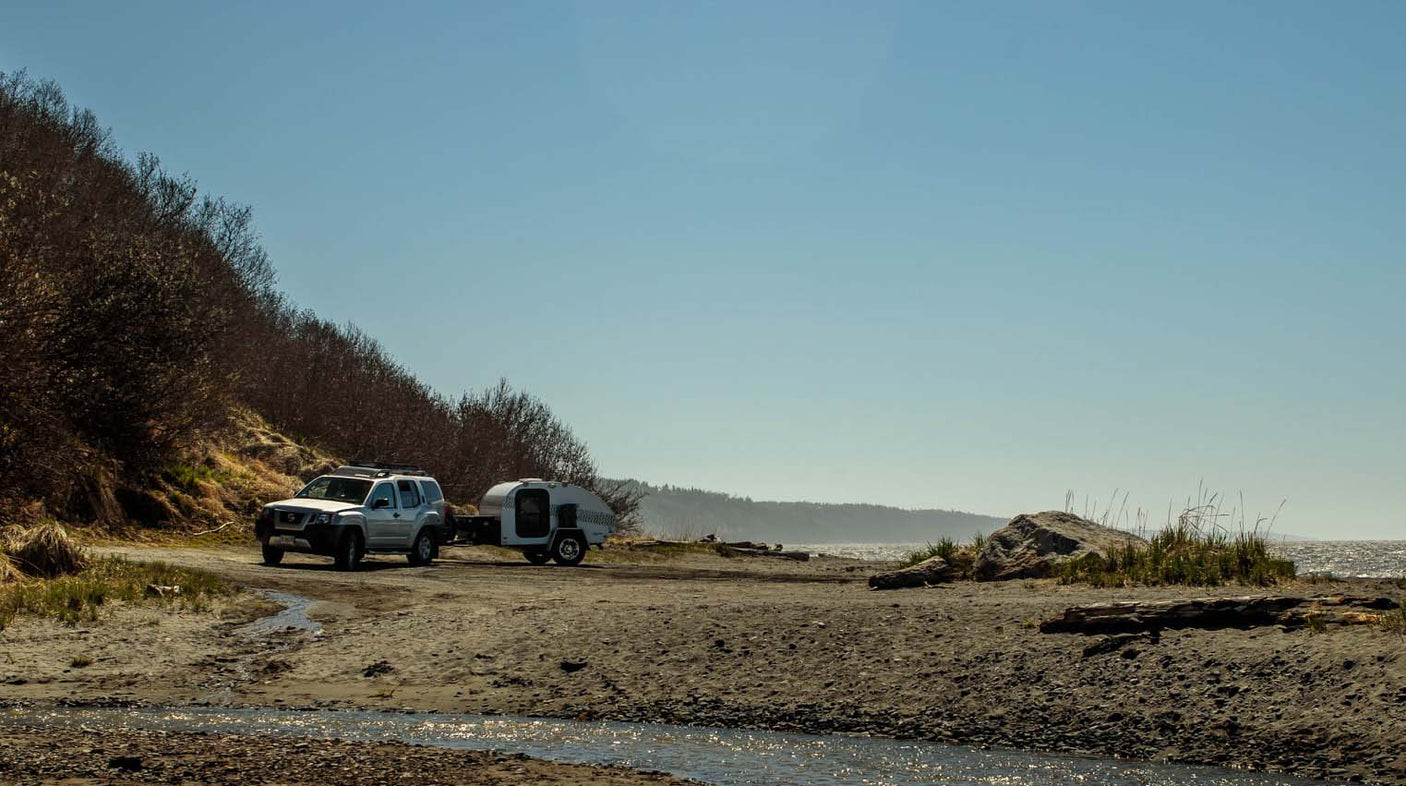 with the abandoned Igloo Hotel in the background.|Silver SUV towing a white RV rental teardrop camper parked on a sandy beach by the ocean.|Compact rv rental teardrop camper features a fully equipped outdoor kitchen with stove
