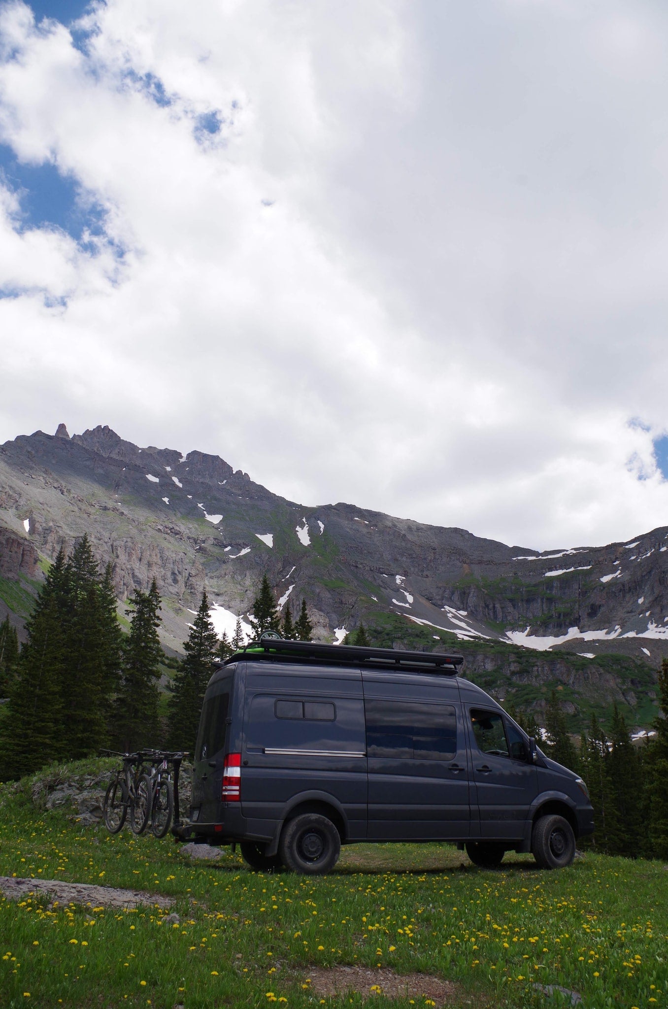 Dark gray RV rental van with bikes, parked in a mountain meadow with yellow flowers & snow-capped mountains. — photo 9