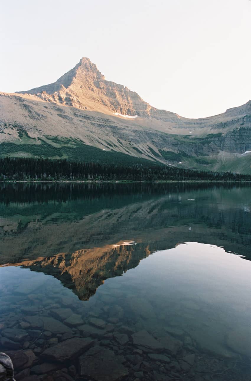 A sharp mountain peak reflects perfectly in a calm, clear lake with visible rocks below. Great RV rental destination!