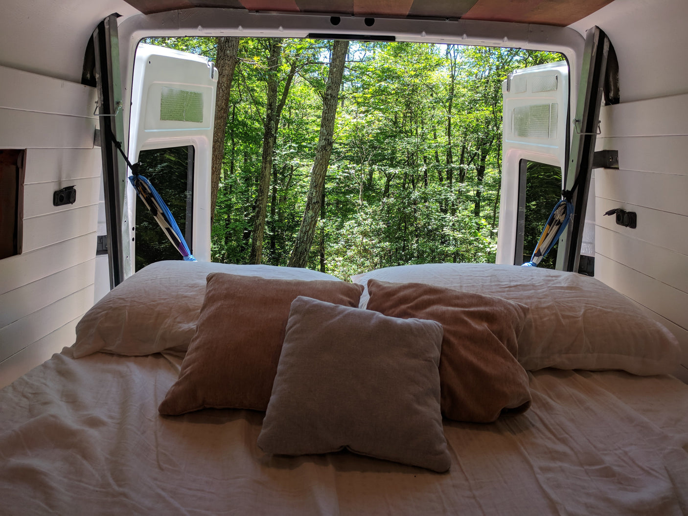 Inside a cozy RV rental: a bed with white sheets and pillows, offering a lush forest view from open rear doors.
