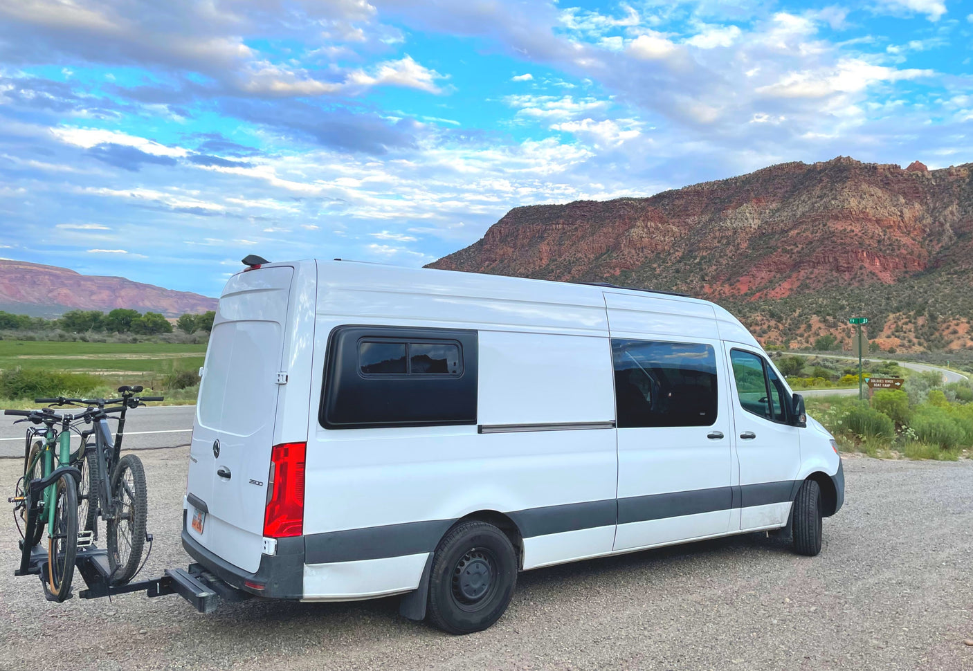 Scenic RV rental with two bikes, parked at a trailhead against a backdrop of red rock mountains and a green valley.