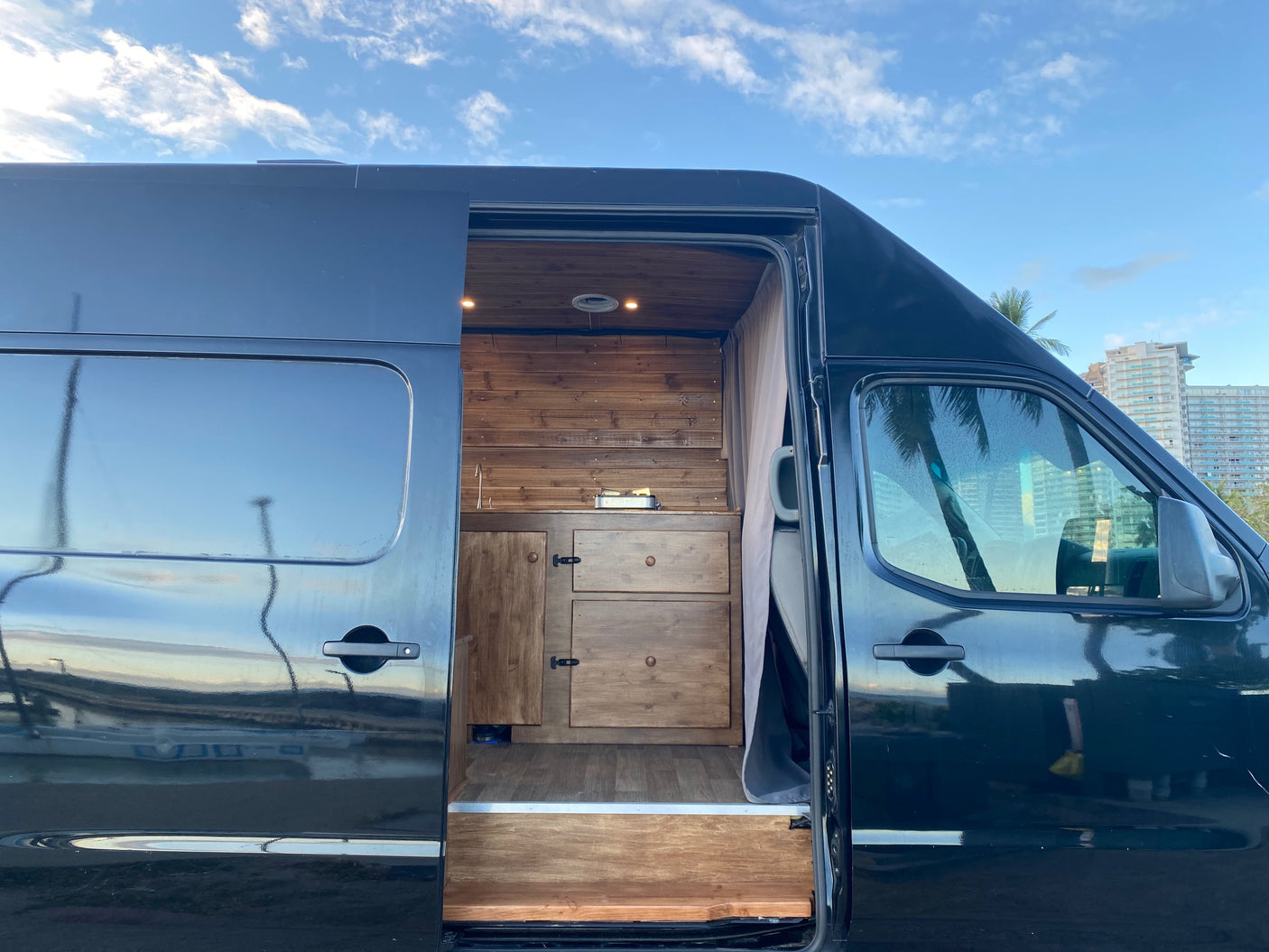 Explore this custom black camper van, perfect as an rv rental. Its open side reveals a cozy wooden kitchen & storage area.