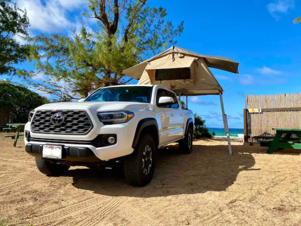 This white Toyota Tacoma truck with a rooftop tent offers a unique RV rental experience at a beautiful beach campsite.