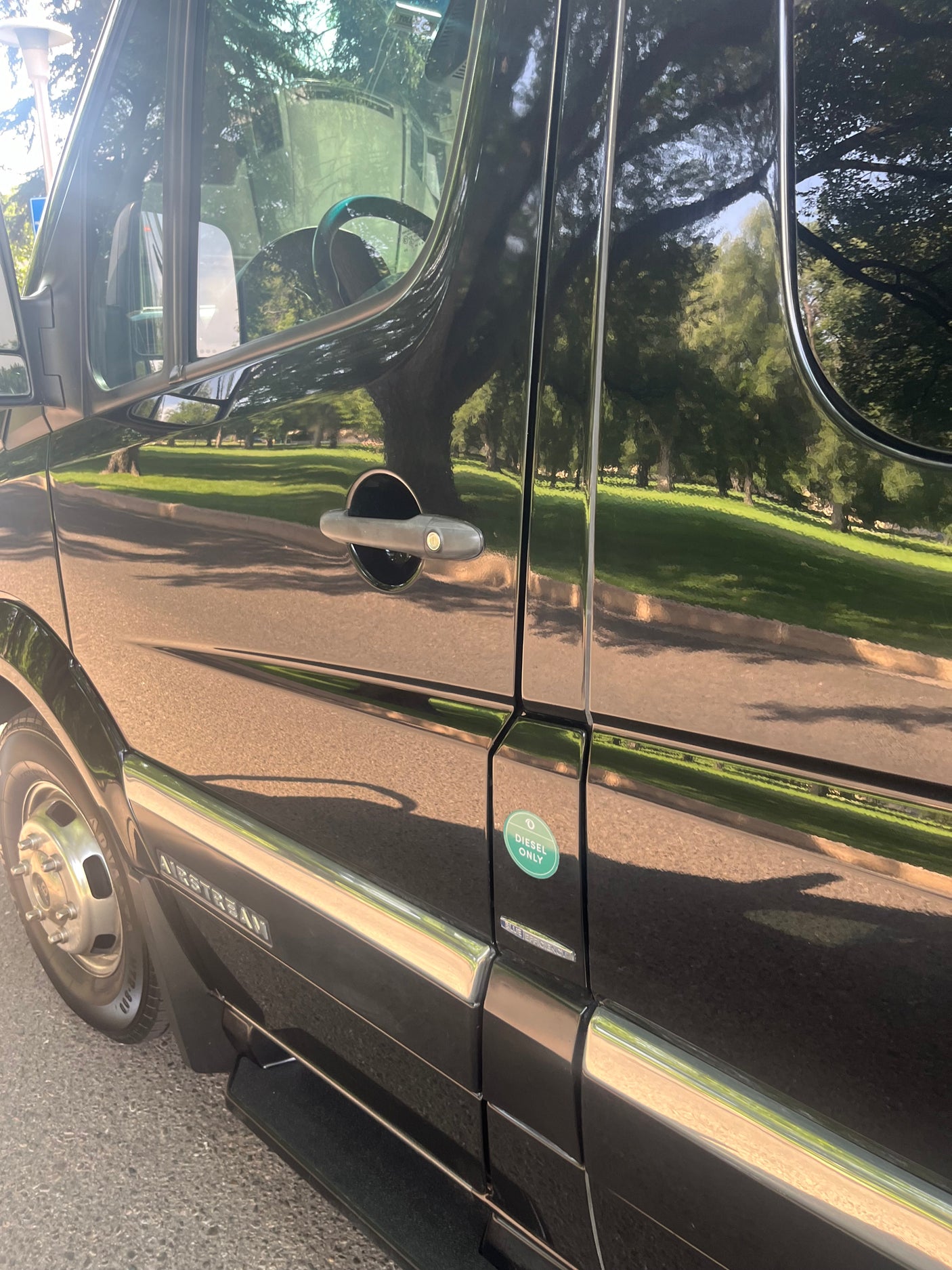 Black Airstream RV rental, a diesel van, parked reflecting sunny trees and grass. Features a "Diesel Only" sticker.