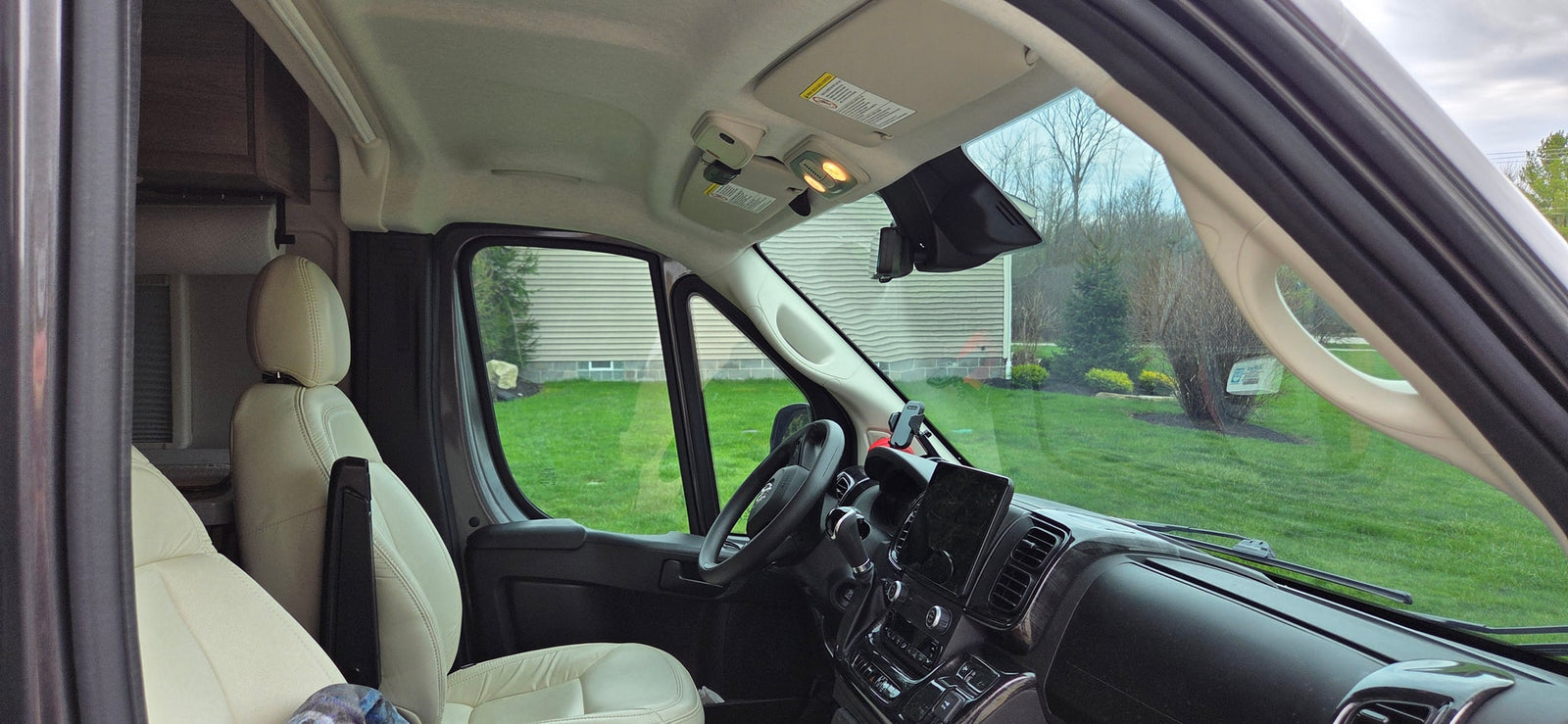 Interior view of an RV rental cockpit featuring light-colored seats, a modern dashboard, and a yard view through the windshield.