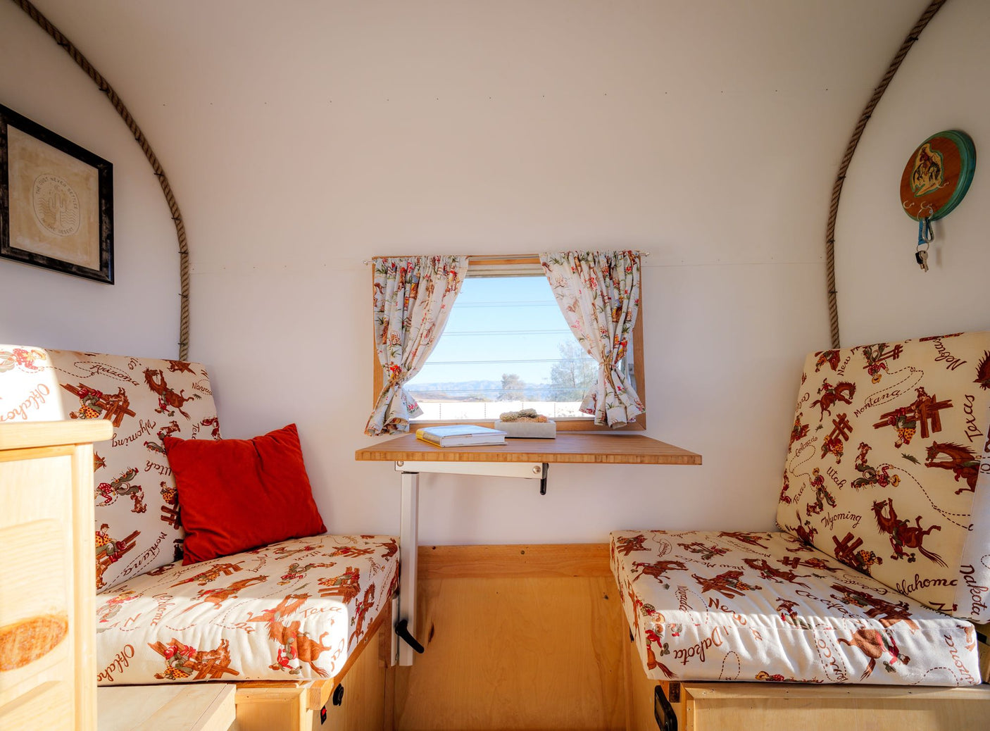 Cozy wagon-style RV rental interior features patterned benches, a fold-down table, window with curtains, and rustic decor.
