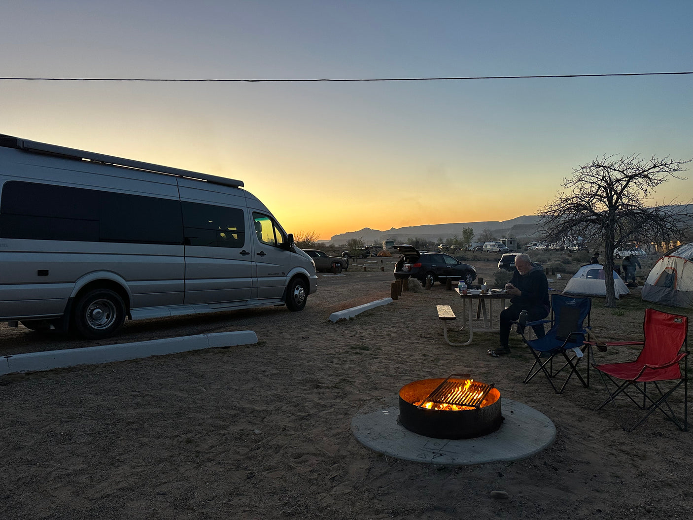 Campsite at sunset: an RV rental, warm campfire, and people enjoying the desert evening amidst tents and vehicles.