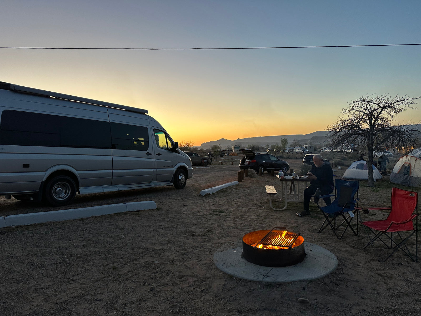 Campsite at sunset: an RV rental, warm campfire, and people enjoying the desert evening amidst tents and vehicles.