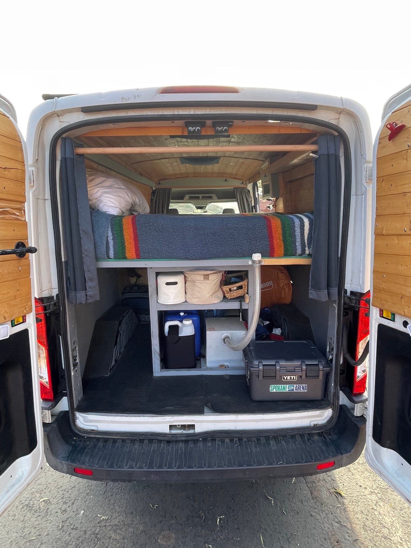 RV rental: Back of a camper van with doors open, revealing a bed, built-in storage with water containers, and a tidy interior.