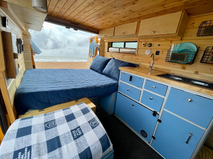 Cozy RV rental interior with a blue bed and kitchen, parked right on a sandy beach overlooking the ocean view.