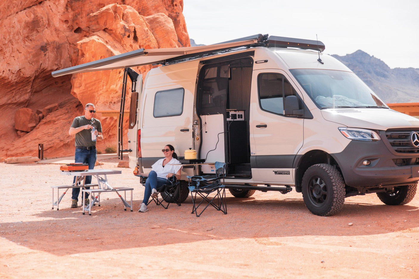 RV rental adventure: A couple enjoys outdoor cooking and relaxation next to their beige camper van in a stunning red rock desert.