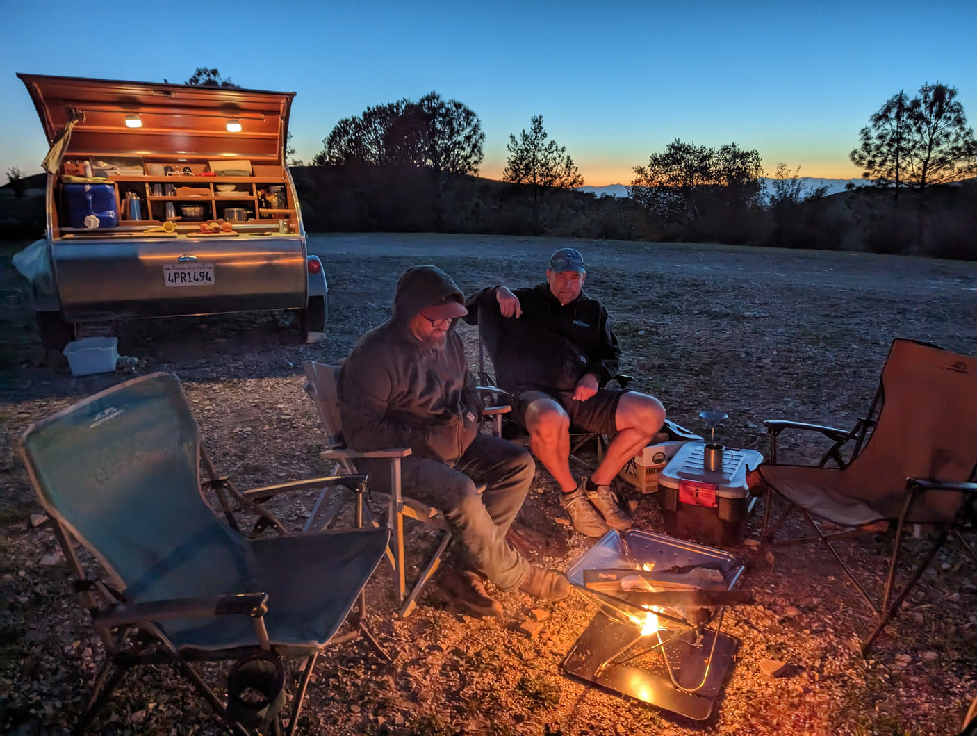 RV rental adventure: Two men by a warm campfire at dusk, next to a detailed, open camper kitchen.