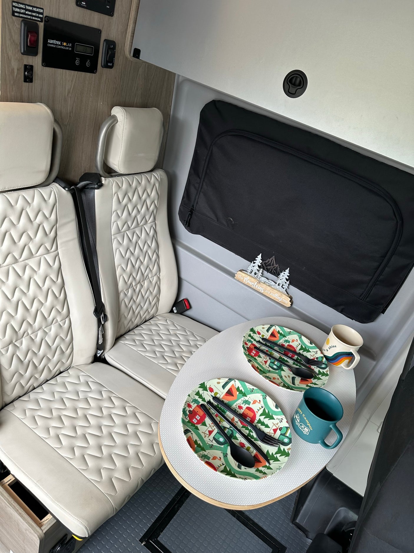 RV rental interior shows cream seats, a dining table set for two, a solar charge controller, and "The Mountains are calling me" decor.
