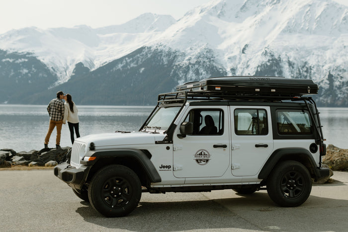 White Jeep RV rental with rooftop tent by an Alaskan lake. Couple enjoys snowy mountain views.