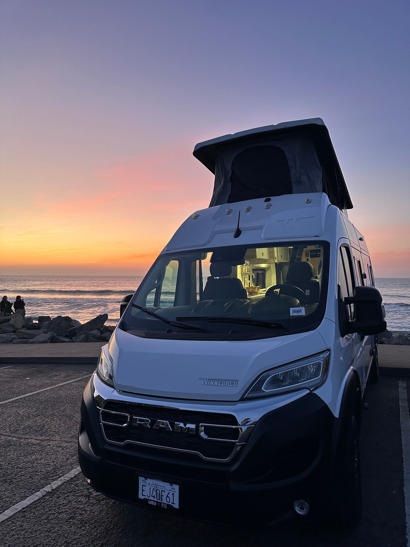 White Winnebago rv rental with pop-top, parked by the ocean at sunset. Interior lights on, ideal for coastal travel.