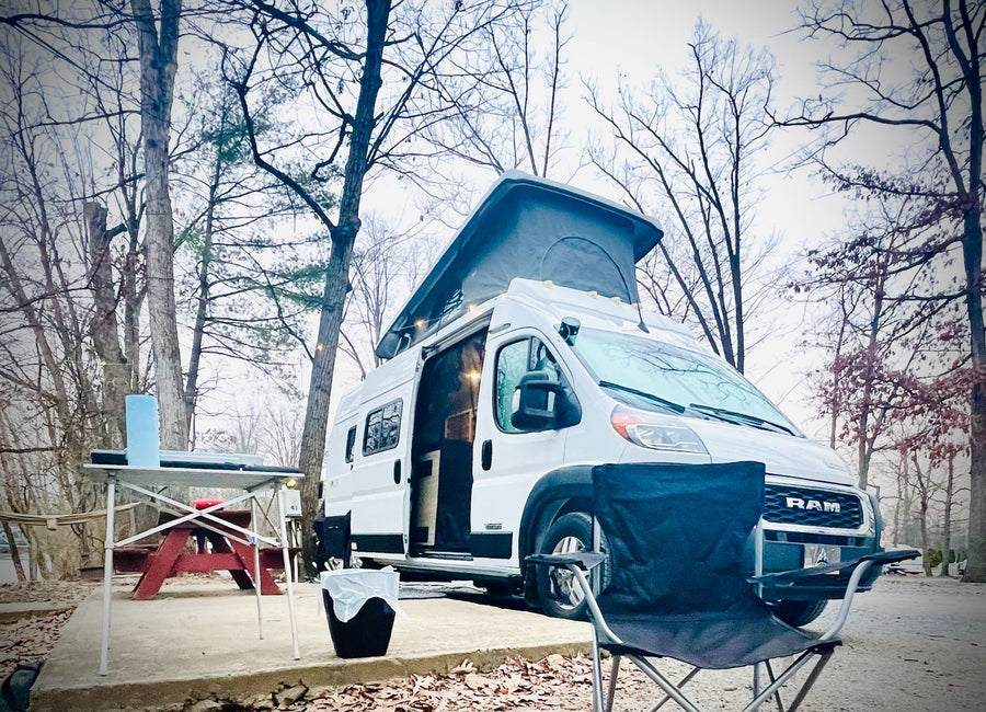 parked at a wooded campsite with a folding table and chair.|White camper van RV rental with open side and front doors