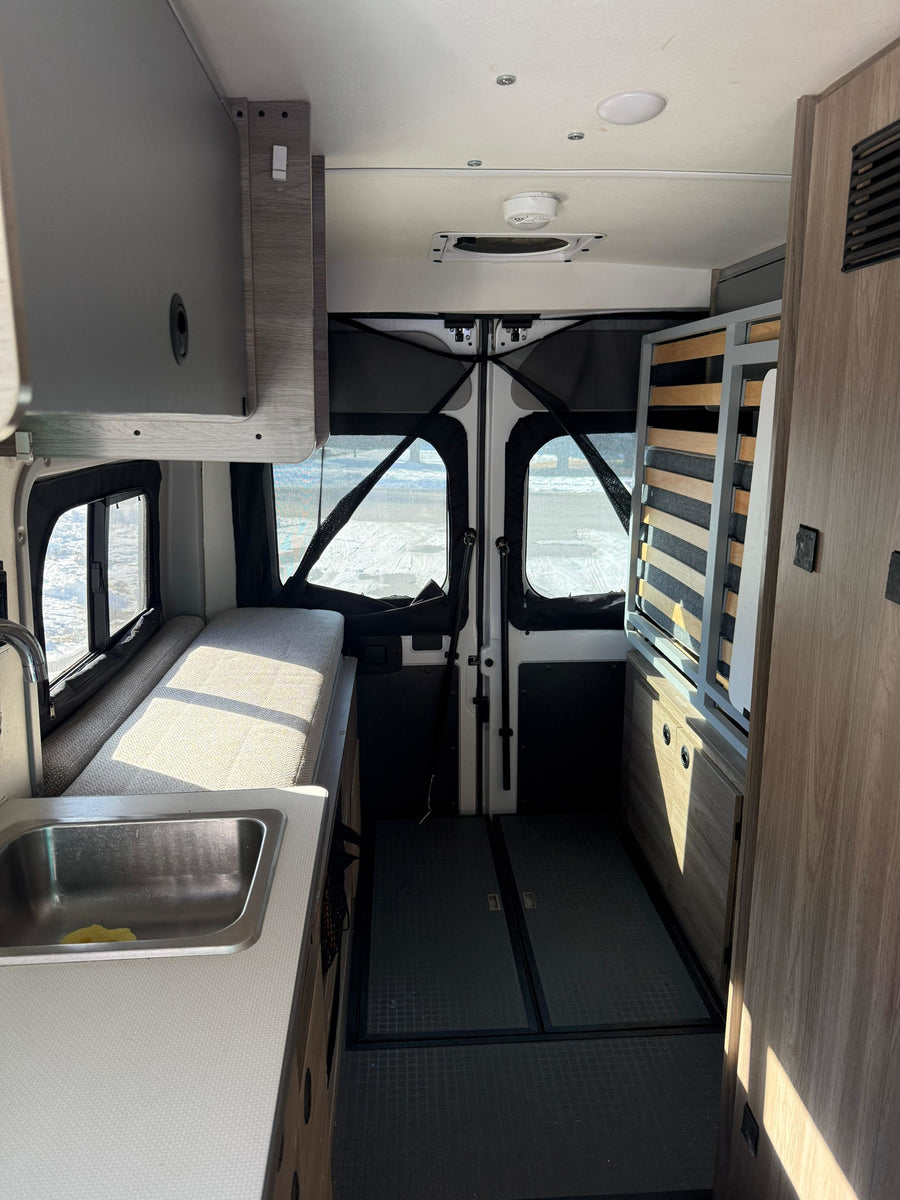 Inside a modern rv rental: kitchen sink, bench bed, overhead cabinets, and rear screens for adventure. Sunlight fills the space.