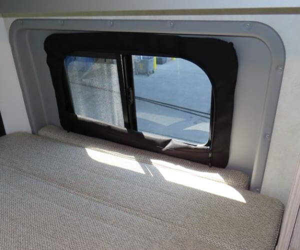 light-colored upholstered bench next to a bright window. A black latch is on the adjacent surface.|Explore in this grey Winnebago Solis pop-top camper van