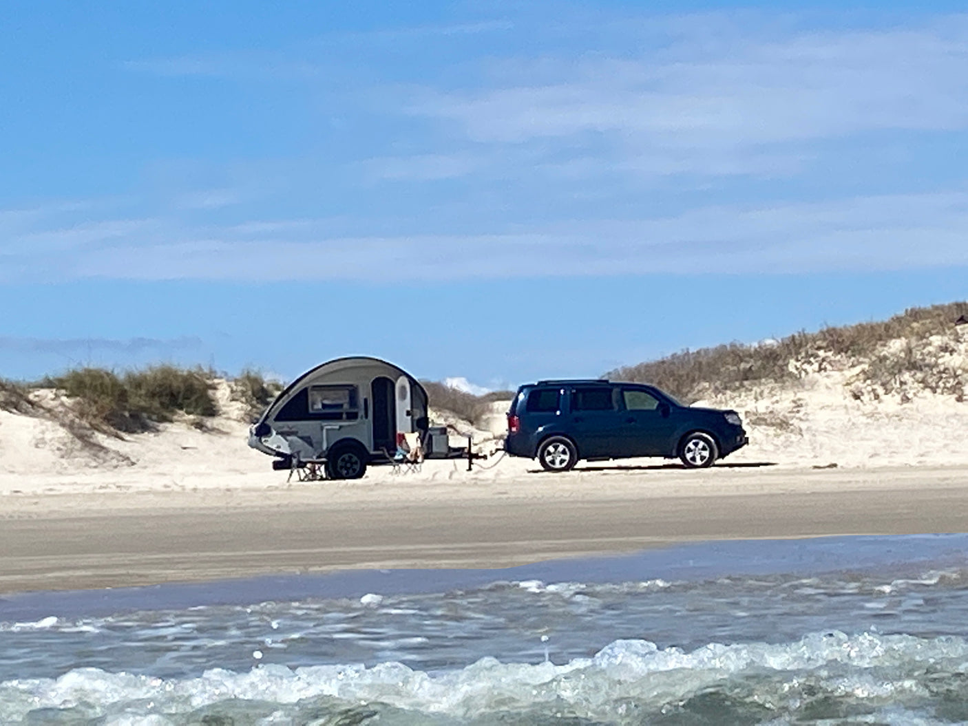 Beach camping with an RV rental! A dark SUV tows a grey teardrop trailer on a sunny sandy beach with ocean waves in the foreground.