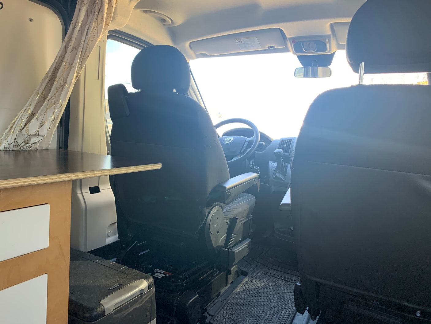RV rental camper van interior from the living area: driver's cockpit, passenger seat, and portable fridge on the left.