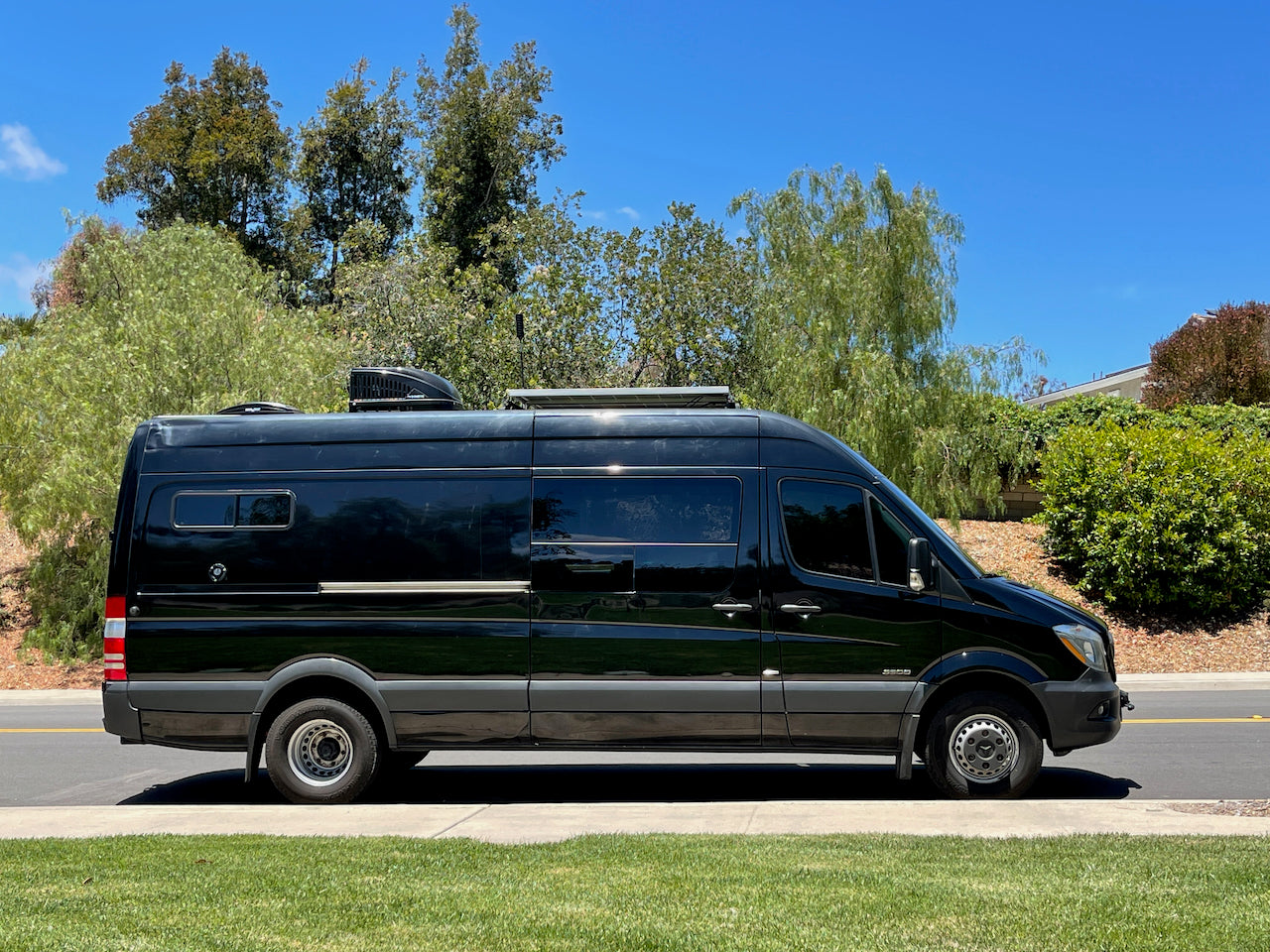 Sleek black Mercedes Sprinter camper van, available as an rv rental, parked on a street with green landscaping.