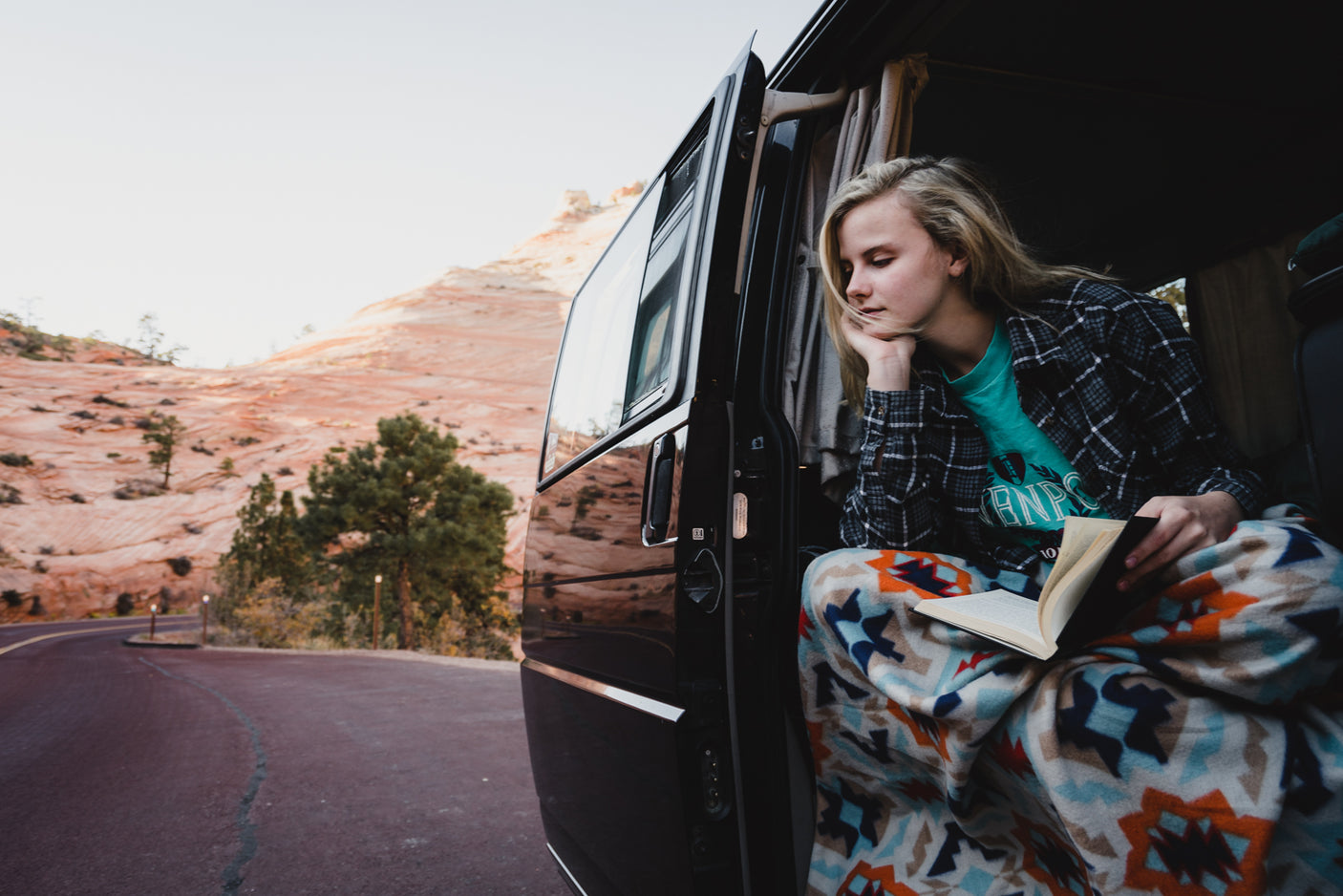 Relaxing RV rental moment: Young woman reading in van door, cozy in a blanket, overlooking scenic desert.