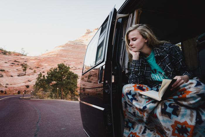 Relaxing RV rental moment: Young woman reading in van door, cozy in a blanket, overlooking scenic desert.