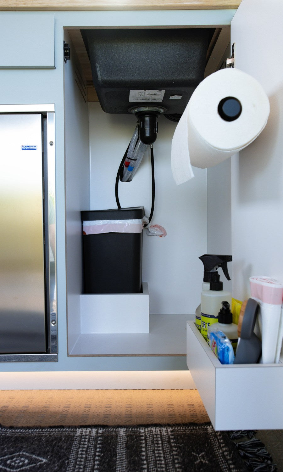 Under-sink area in an RV rental, showing a trash can, paper towels, cleaning supplies, and a compact fridge. Optimized storage.