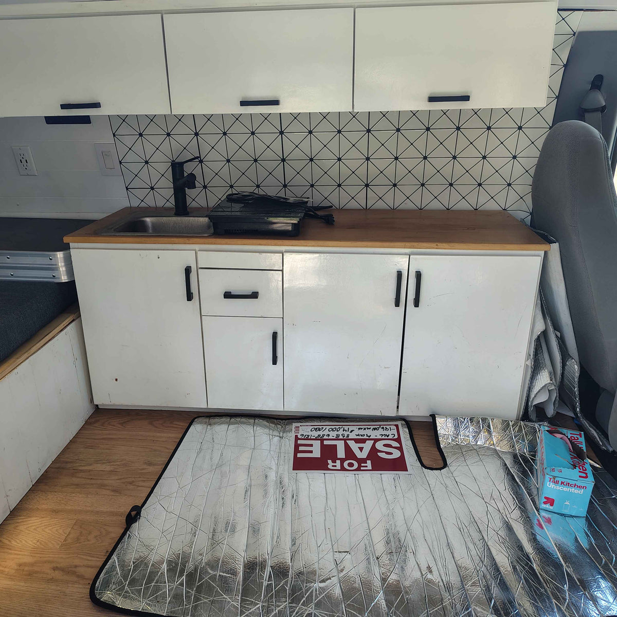 A compact RV rental kitchen features white cabinets, wood counter, sink, black faucet, and a geometric backsplash. A "FOR SALE" sign is on a sunshade. — photo 8