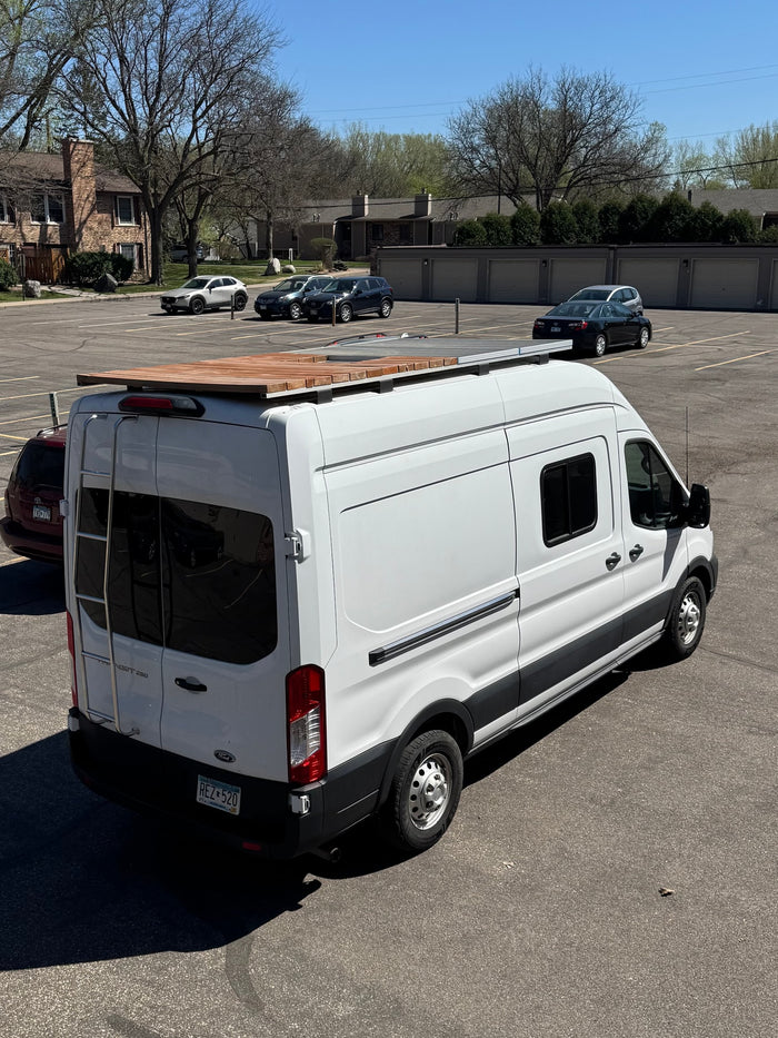 A white Ford Transit camper van, equipped with a roof deck and solar panels, visible in a parking lot. Ideal for rv rental adventures.