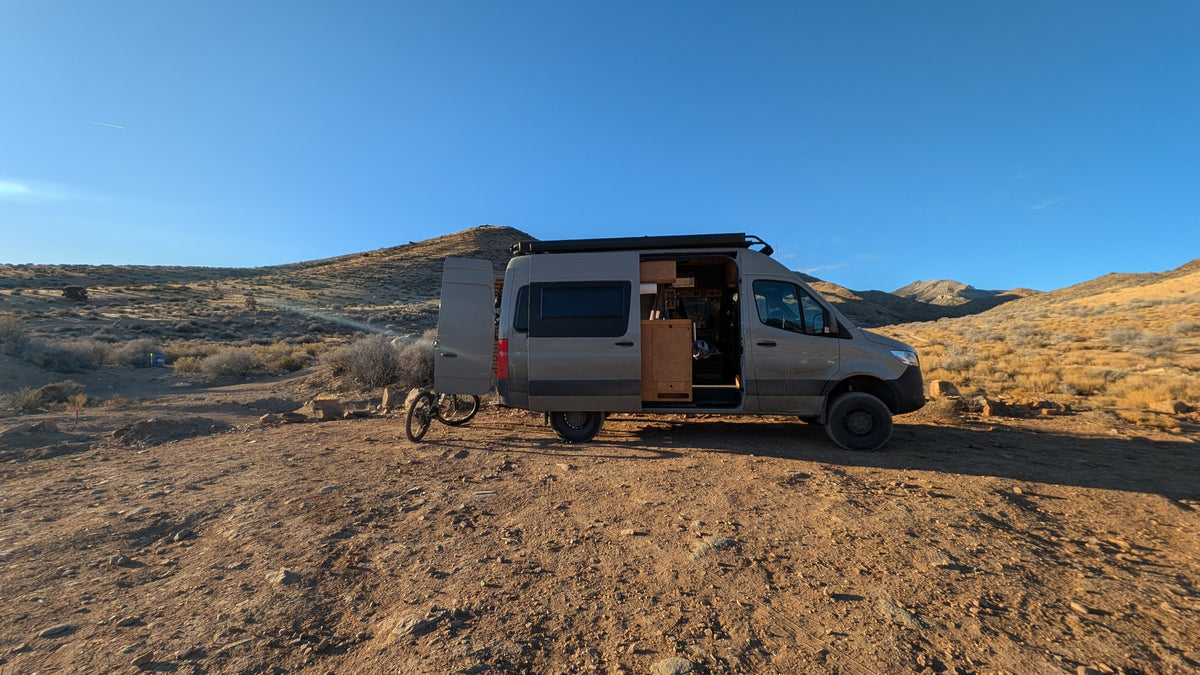 Gray camper van for RV rental, parked on dirt in a sunny, hilly desert. Doors open, bike attached, ready for off-grid van life adventures.