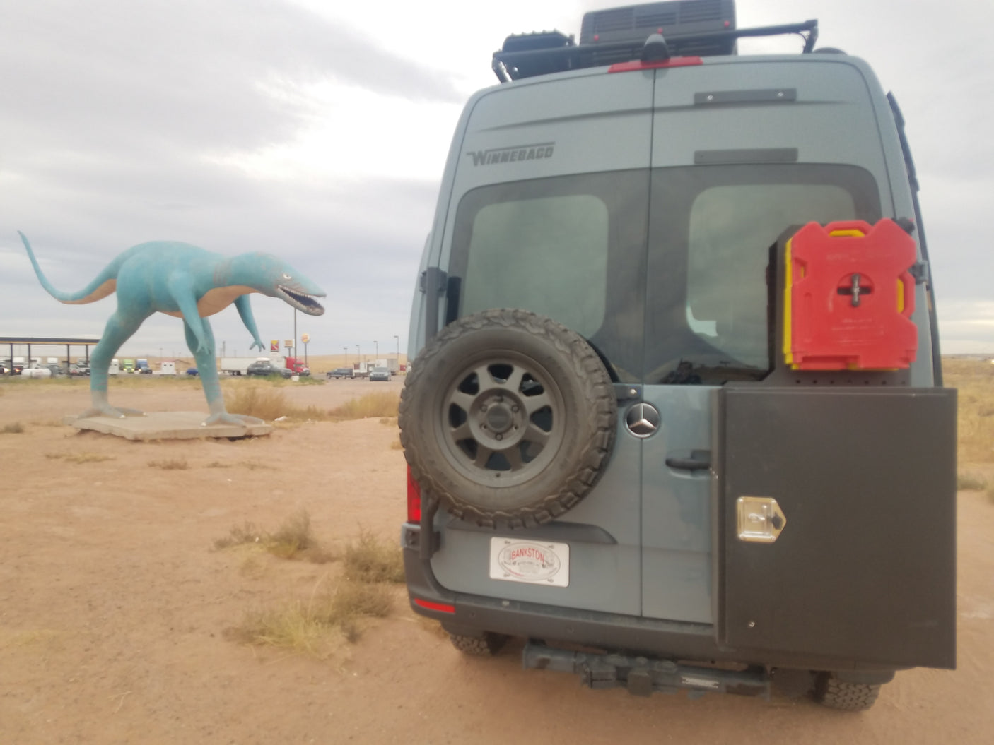 This rv rental Winnebago camper van with spare tire, storage & fuel sits by a large blue dinosaur statue. Roadside attraction.