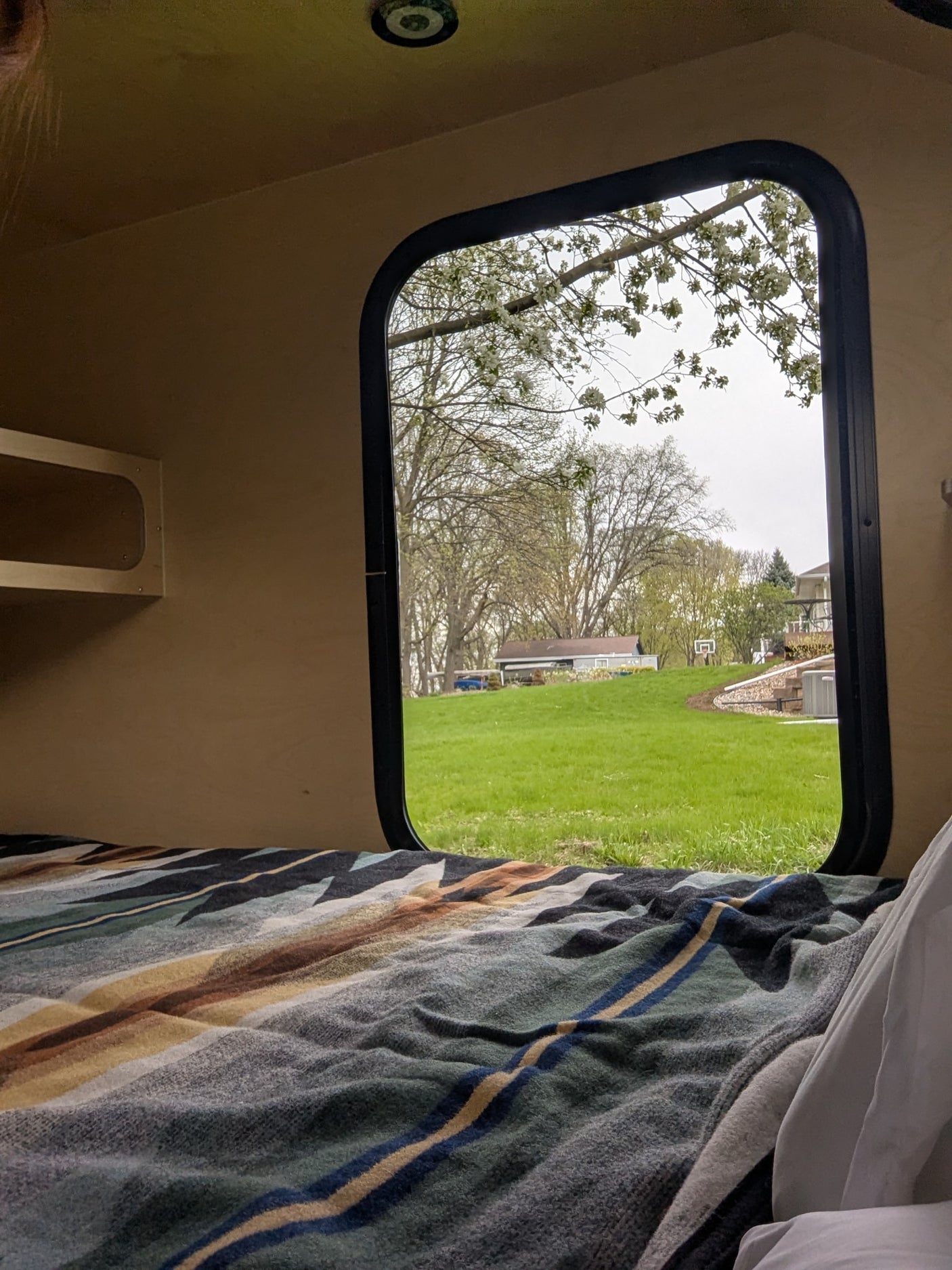 Cozy RV rental interior with a view from the bed: green lawn, blooming trees, and houses outside a rounded window.