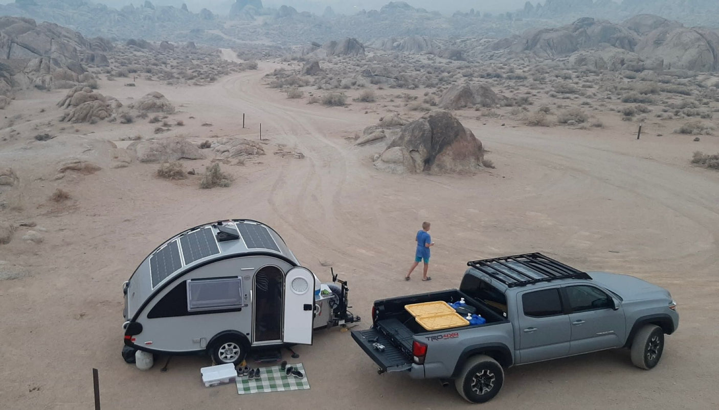 RV rental campsite: Grey pickup, teardrop camper with solar panels (door open, mat), and person in rocky desert.