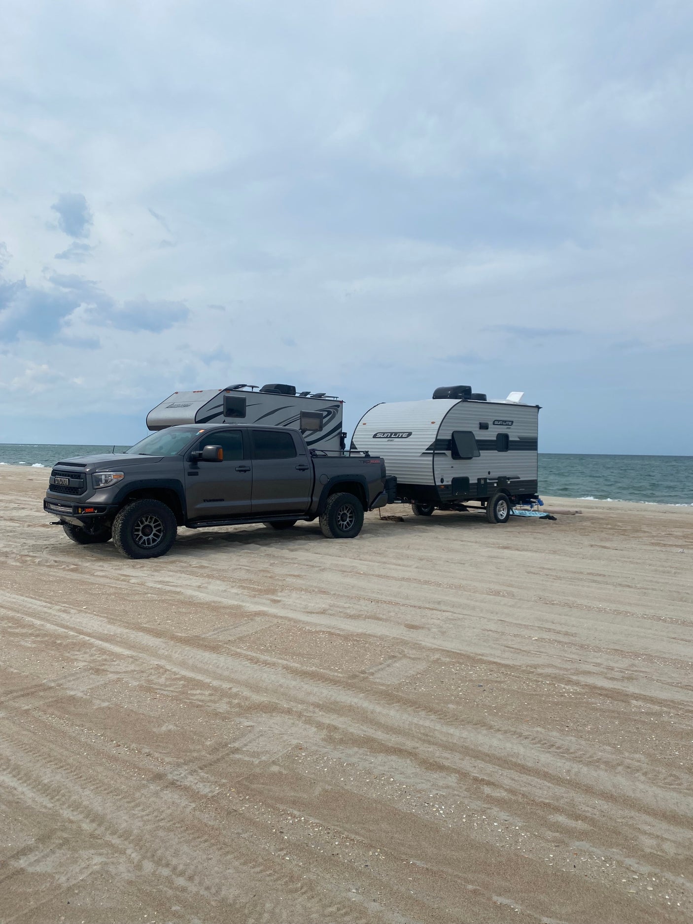 Two RVs, including a Sun Lite Sport towed by a truck, parked on a sandy beach by the ocean. Ideal for an RV rental vacation!