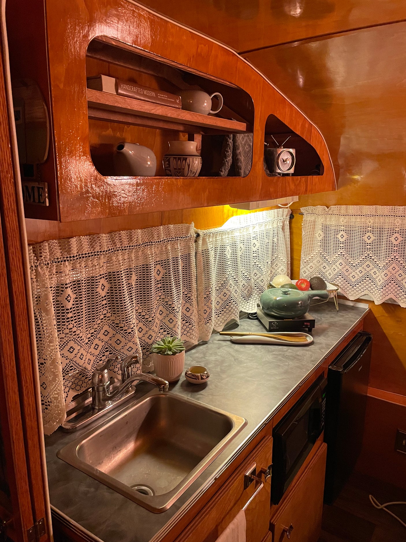 A well-appointed rv rental kitchen featuring rich wood cabinetry, a sink, microwave, induction cooktop, and lace curtains.