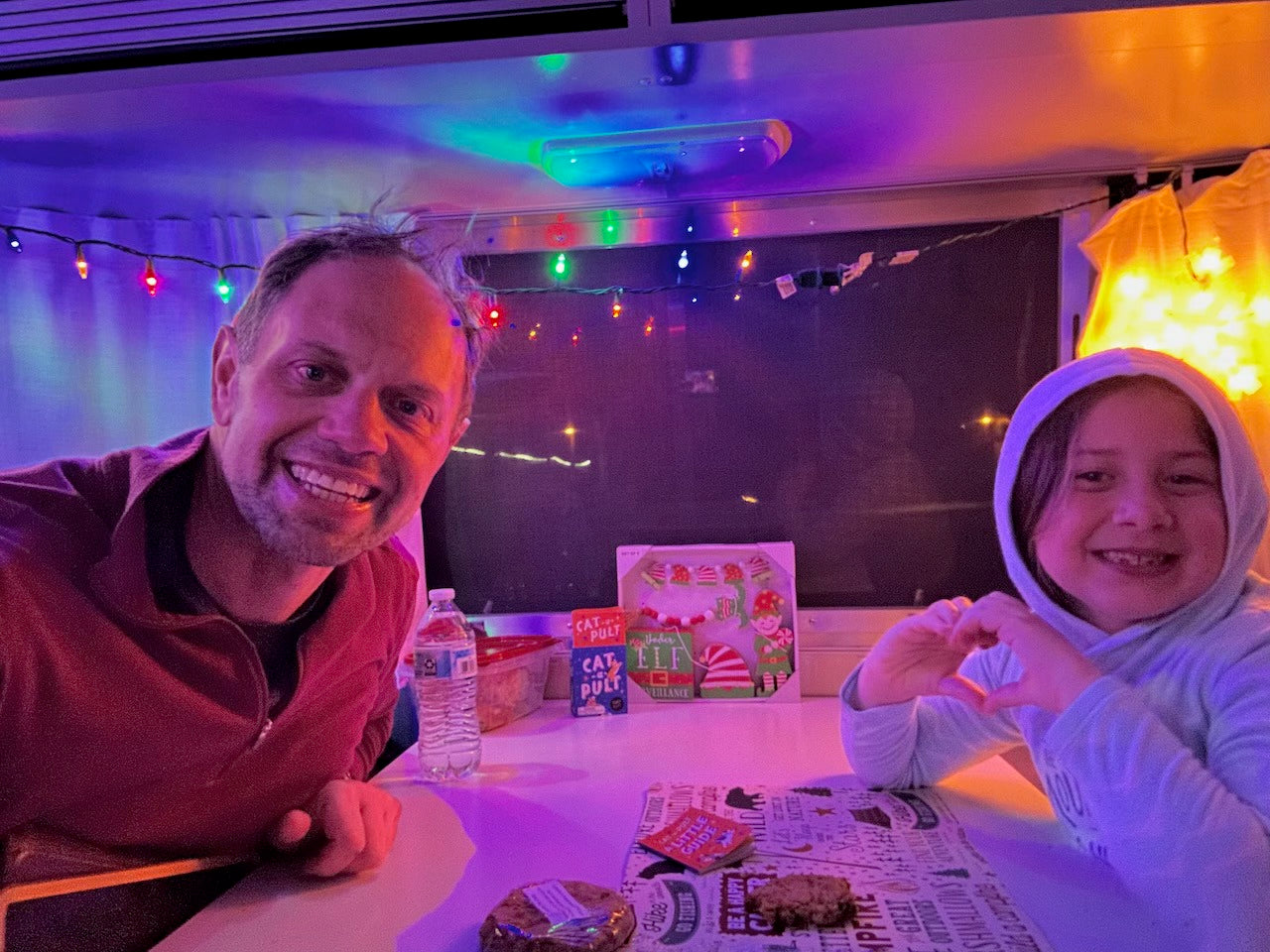 Cozy family fun in an RV rental! Dad and daughter smile, surrounded by colorful lights and treats.